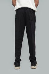 Back view of a model wearing full-length solid black casual trousers featuring a rear patch pocket, comfortable relaxed fit, paired with an untucked white shirt and black athletic sneakers on a plain neutral studio background.