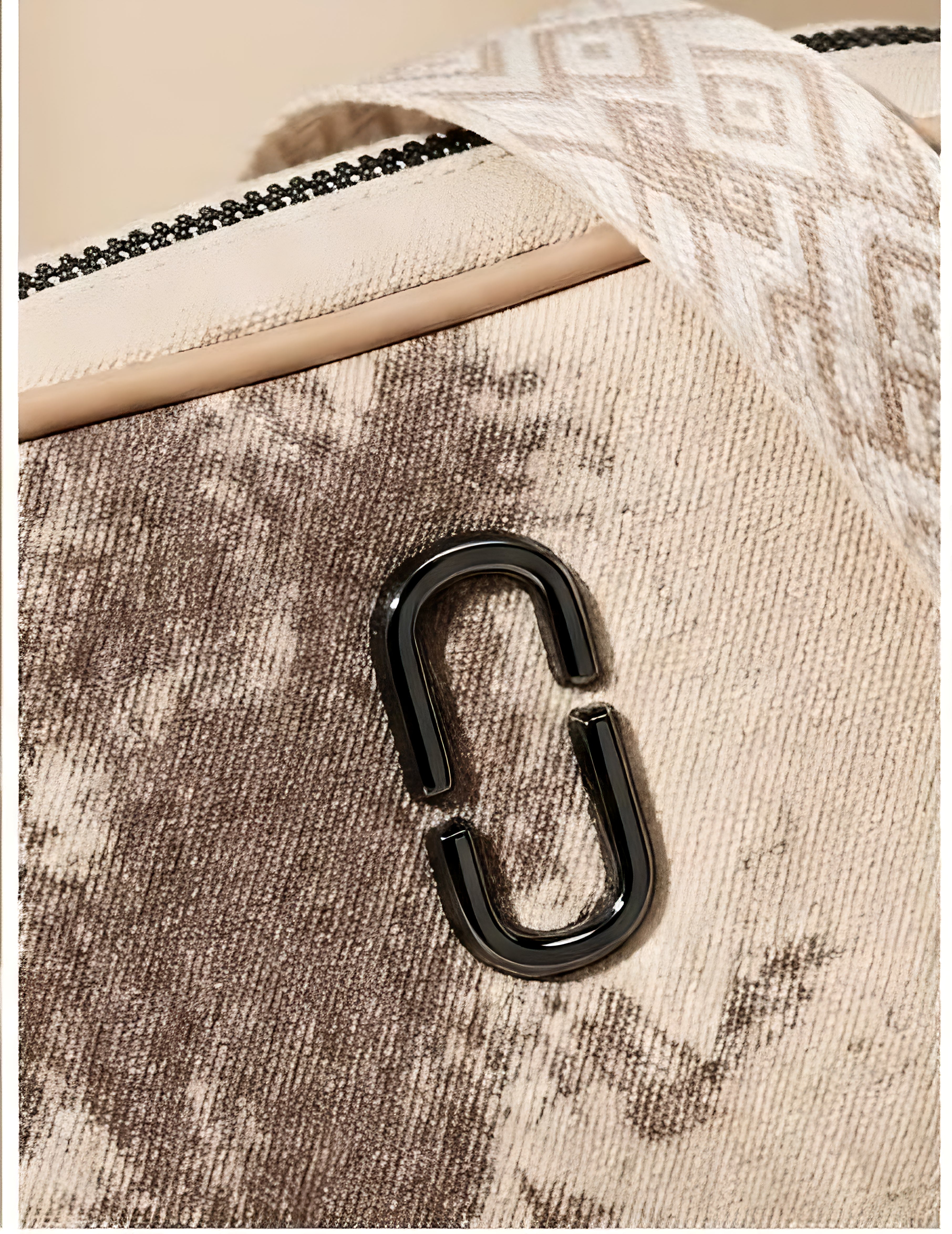 Close-up of a textured canvas bag in beige and brown tones, featuring a glossy black metal C-shaped hardware accent and a patterned cream strap.