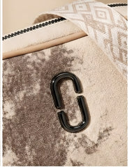 Close-up of a textured canvas bag in beige and brown tones, featuring a glossy black metal C-shaped hardware accent and a patterned cream strap.