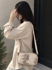 A woman wearing a cream-colored linen shirt and light blue jeans carries a matching cream crossbody bag with a distressed gray pattern and a silver metal O-ring closure. She is standing next to a dark window frame.