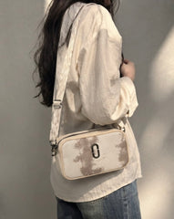 A woman wearing a cream-colored crinkled blouse and blue jeans carries a cream crossbody bag with a brown abstract pattern and a silver metal clasp against a neutral wall.