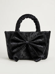 Front view of a black crushed velvet tote handbag featuring a large, decorative bow accent and ruched top handles against a light gray background.
