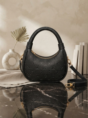 Front view of a black embossed Louis Vuitton Loop hobo handbag on a marble surface with a dried palm leaf vase and books in the background.