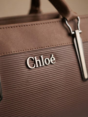 A close-up view of the "Chloé" logo in silver letters on a brown textured handbag.