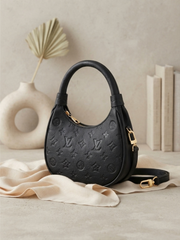 Three-quarter front view of the black Monogram Empreinte Louis Vuitton Loop hobo bag, resting on a beige fabric against a neutral background with a palm fan vase and books.