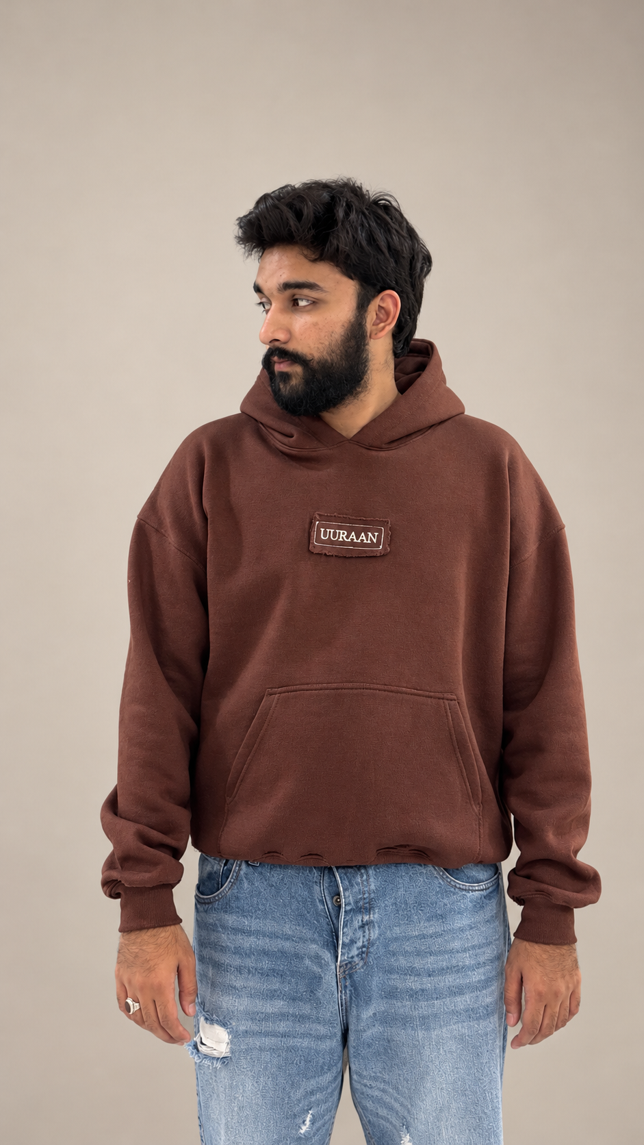 A male model looking to the side, wearing a premium brown oversized fleece hoodie featuring a UURAAN logo patch on the chest and a kangaroo pocket, paired with light blue distressed jeans.