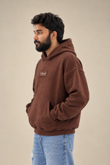 Side profile view of a male model wearing a premium brown oversized fleece hoodie with a UURAAN chest logo patch and hands in front pocket, paired with light blue jeans.