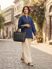 A model wearing a blue embroidered tunic and cream trousers walking down a historic street while carrying the black monogram tote bag.