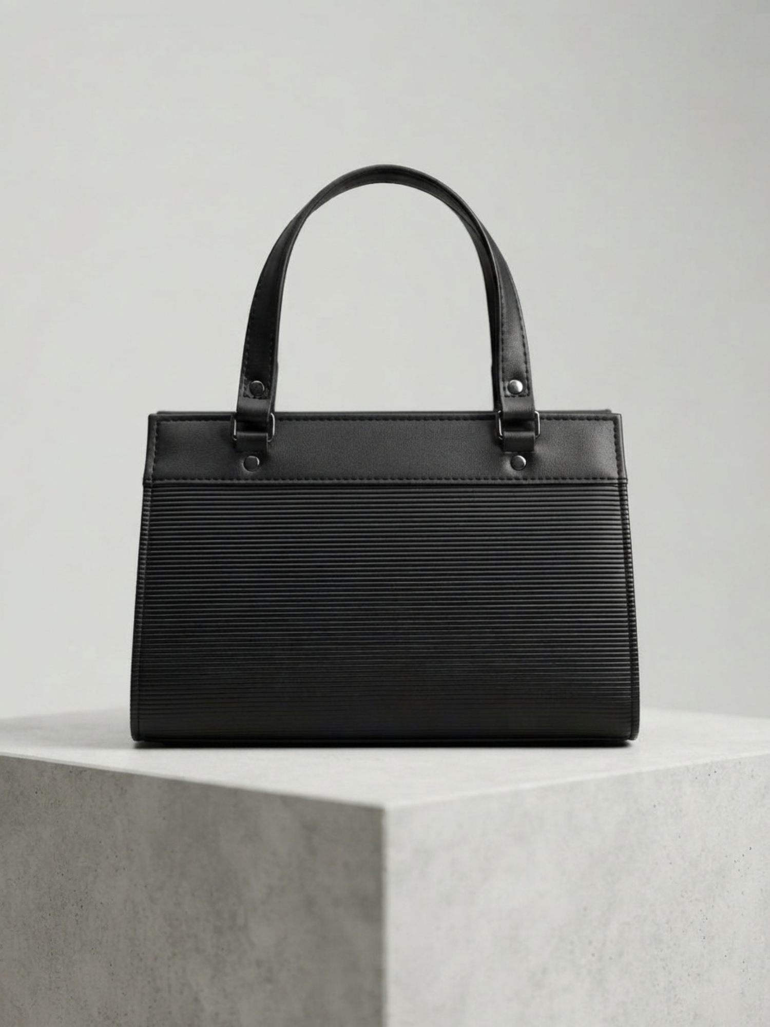Back view of the black textured handbag on a light gray pedestal.