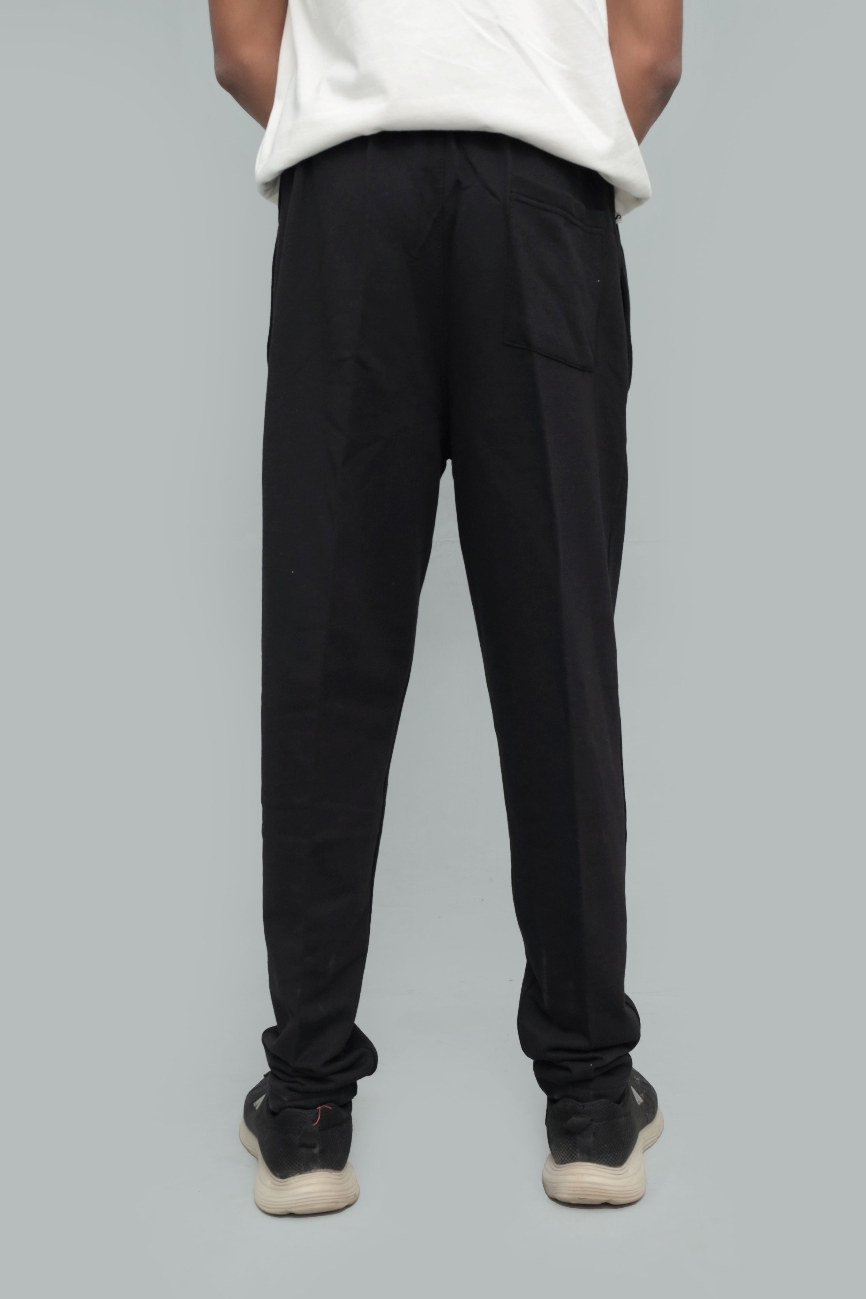 Back view of a model wearing full-length solid black casual trousers featuring a rear patch pocket, comfortable relaxed fit, paired with an untucked white shirt and black athletic sneakers on a plain neutral studio background.