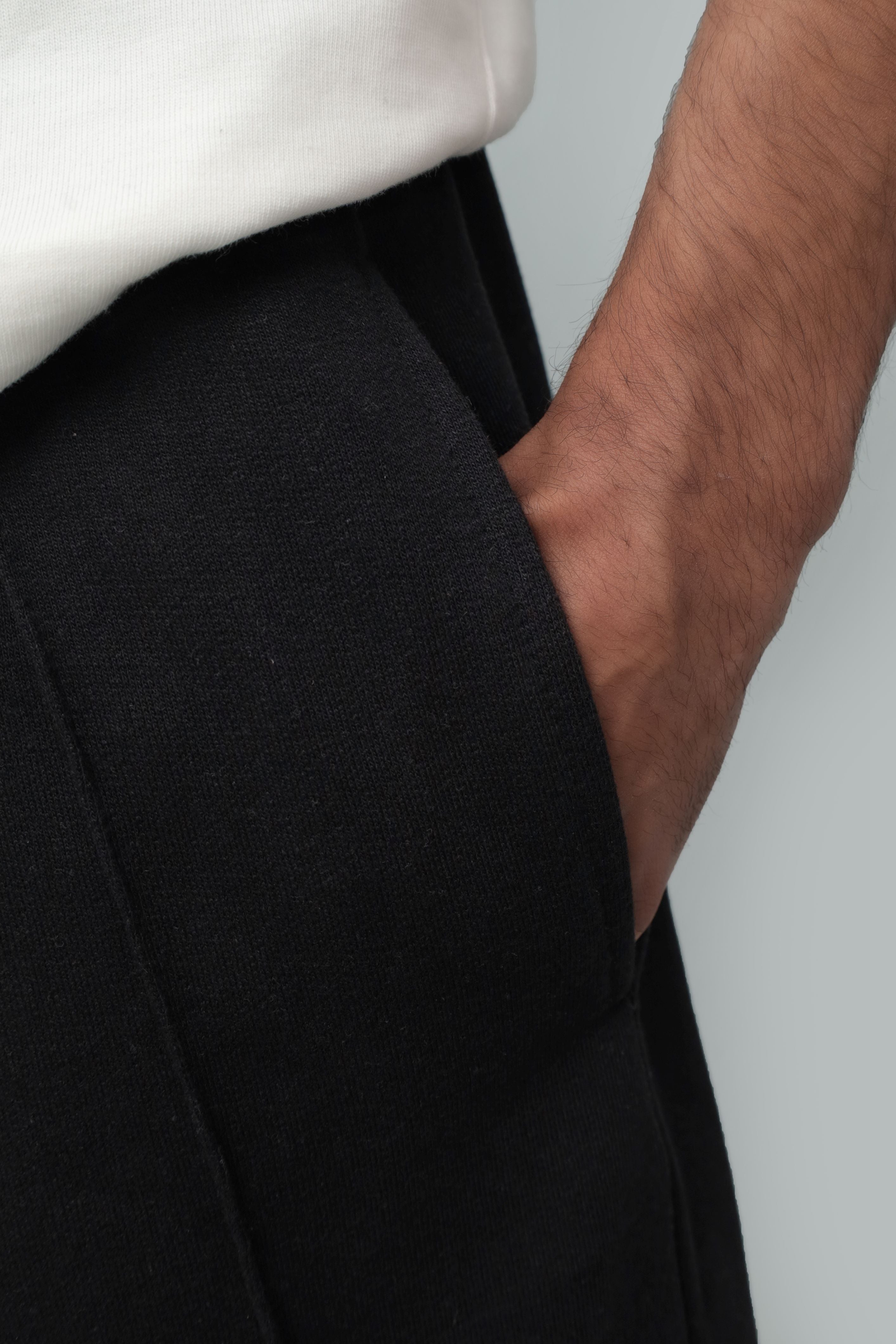 Close-up detail shot of a model's hand resting inside the side pocket of black fleece-lined winter trousers, highlighting the fabric texture, side seam stitching, and elasticated waistband worn under a white top.