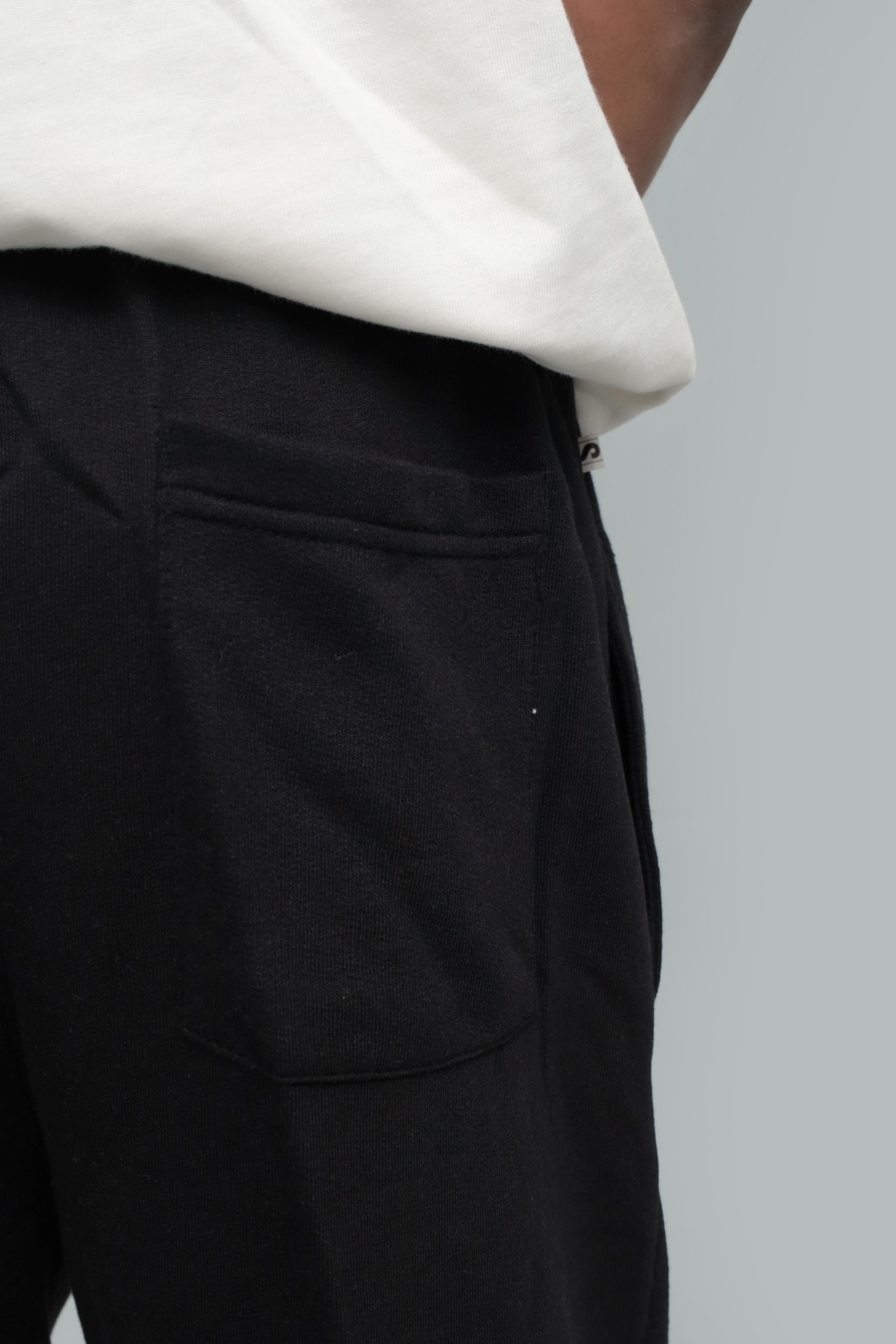 Close-up detail shot showing the back patch pocket, waistband stitching, and fabric texture of black winter trousers, worn with a white top against a light grey background.