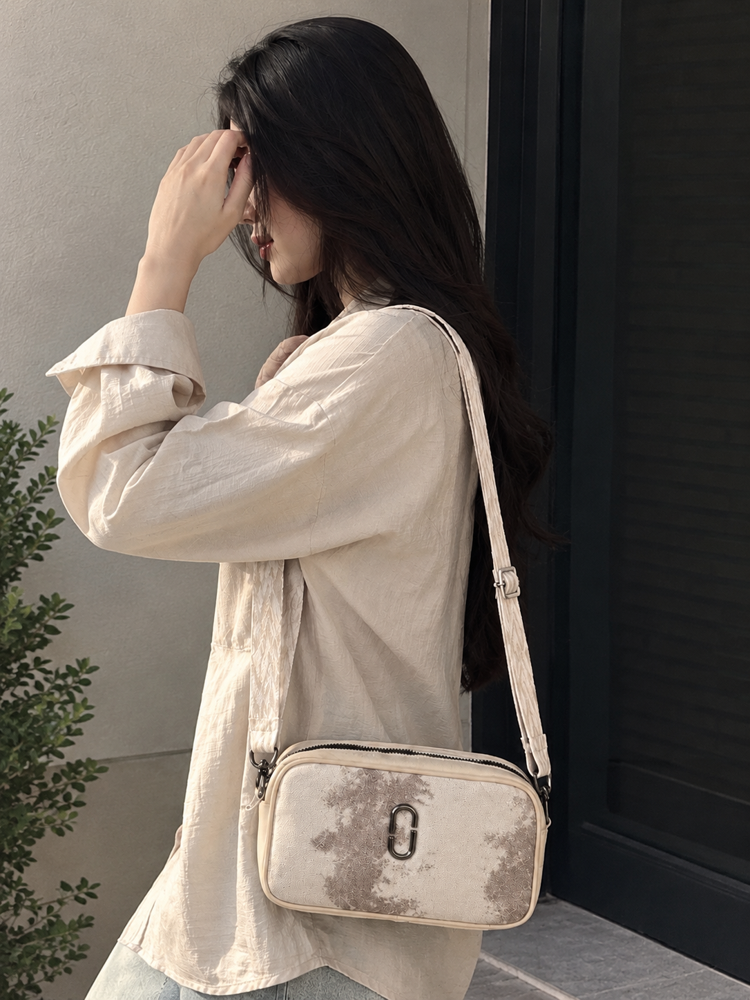 A woman wearing a cream-colored linen shirt and light blue jeans carries a matching cream crossbody bag with a distressed gray pattern and a silver metal O-ring closure. She is standing next to a dark window frame.
