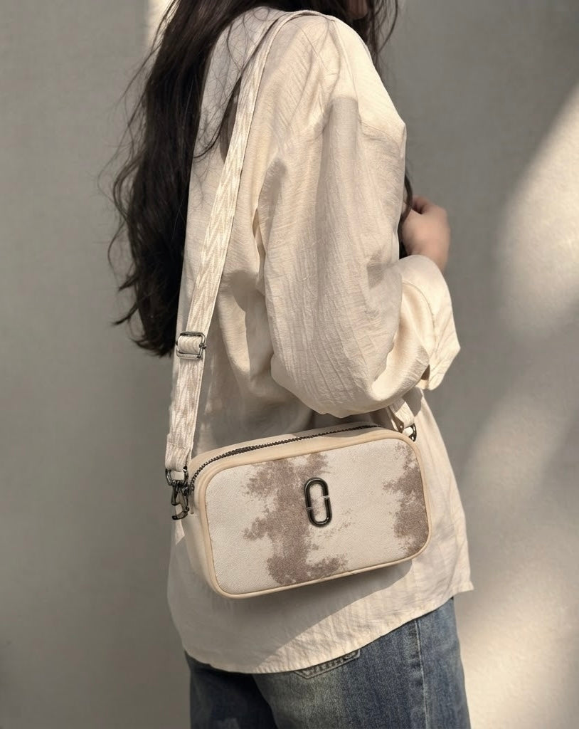 A woman wearing a cream-colored crinkled blouse and blue jeans carries a cream crossbody bag with a brown abstract pattern and a silver metal clasp against a neutral wall.