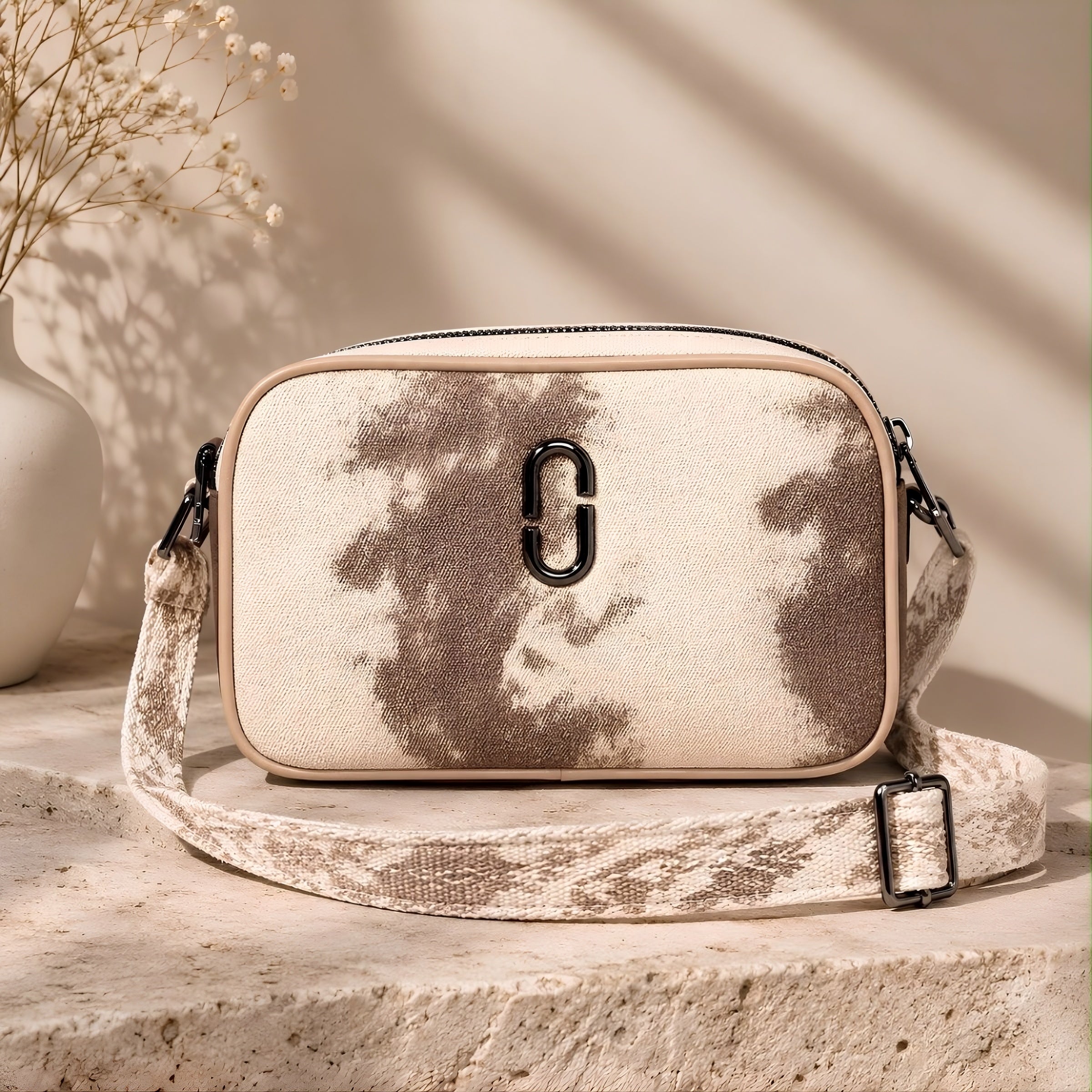 A wrectangular bag with a beige and brown tie-dye pattern, featuring a prominent black metal 'C' logo on the front and a matching patterned shoulder strap, displayed on a stone pedestal with a vase of dried flowers in the background.