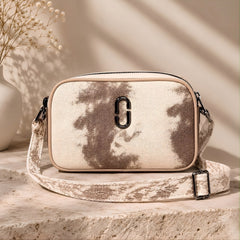 A wrectangular bag with a beige and brown tie-dye pattern, featuring a prominent black metal 'C' logo on the front and a matching patterned shoulder strap, displayed on a stone pedestal with a vase of dried flowers in the background.