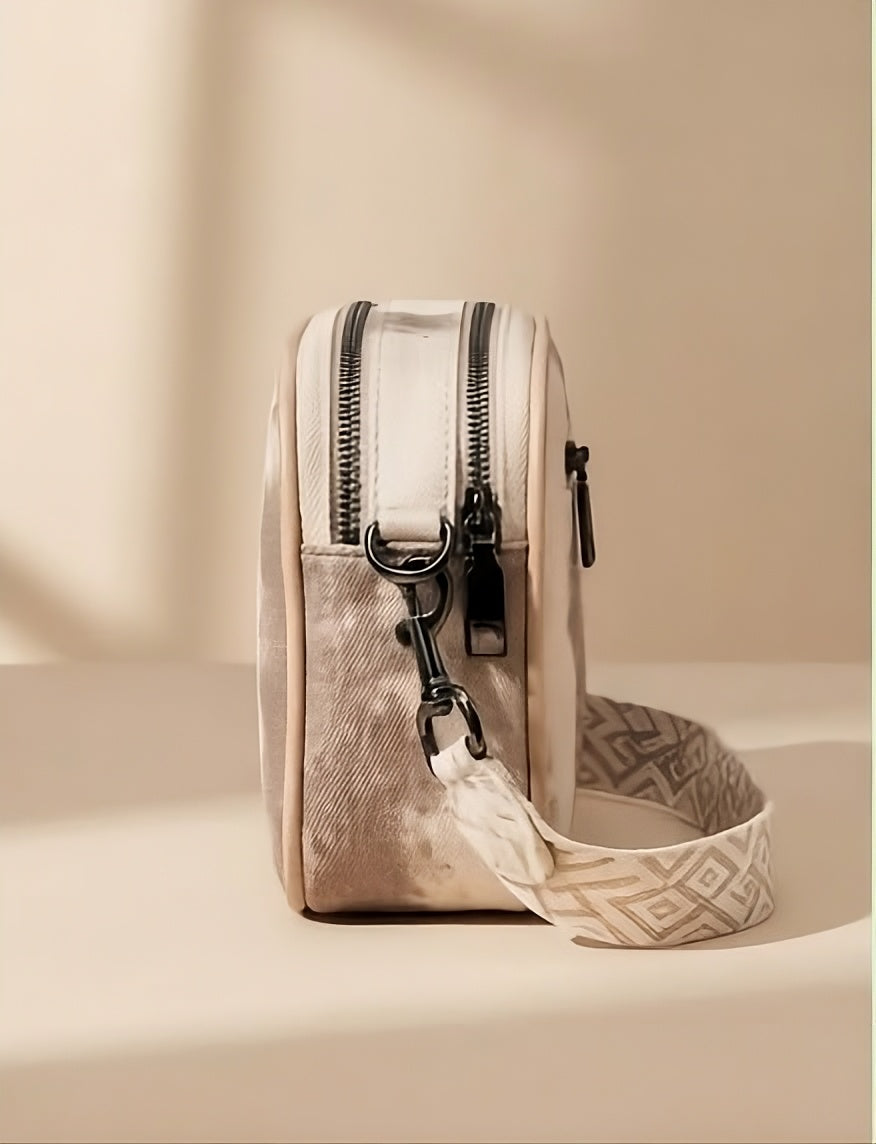 A small beige crossbody bag with dual black zippers, black hardware, and a patterned beige-and-white strap, photographed on a warm beige surface with soft shadows.