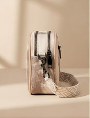 A small beige crossbody bag with dual black zippers, black hardware, and a patterned beige-and-white strap, photographed on a warm beige surface with soft shadows.