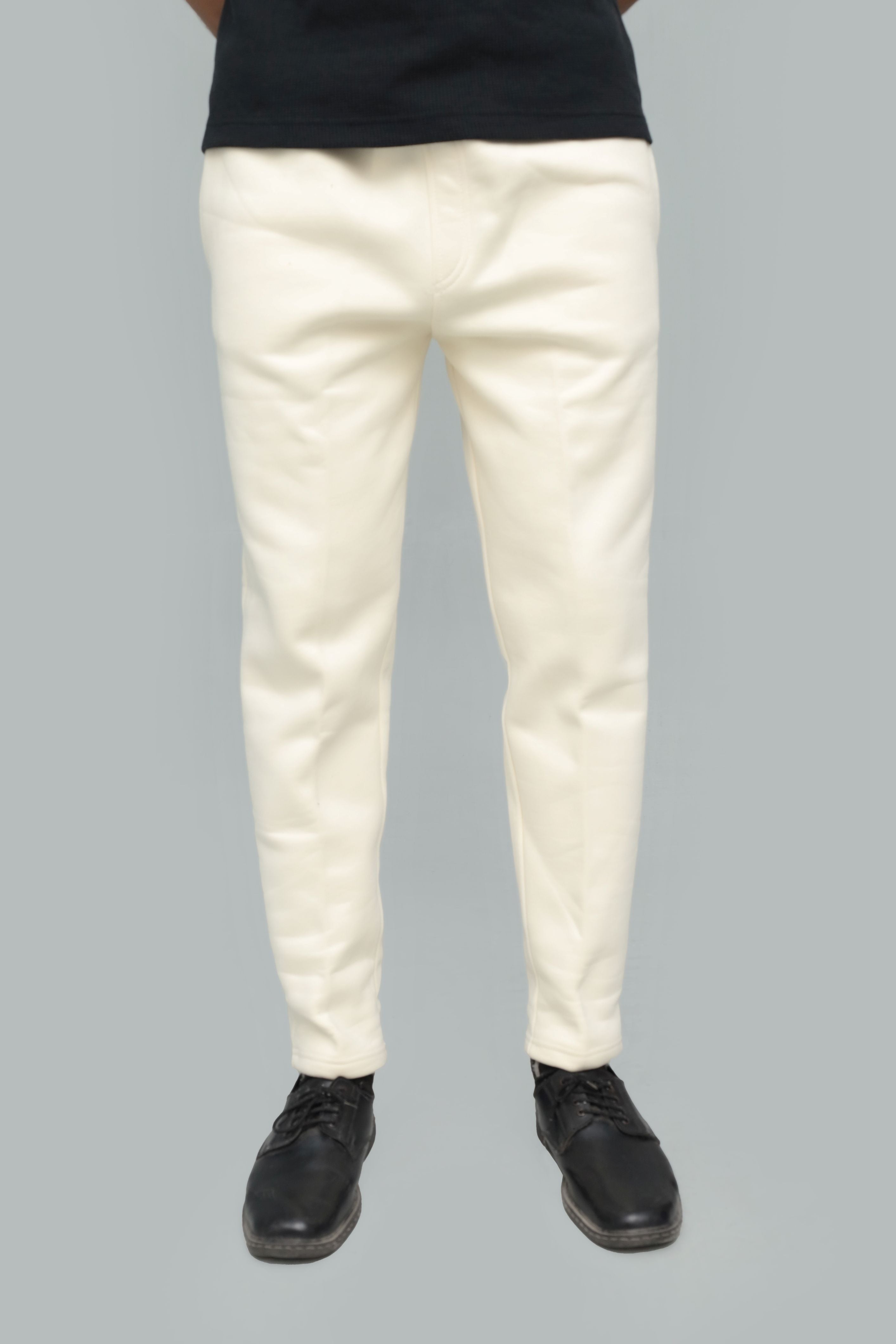 A person shown from the waist down wearing off-white slim-fit trousers, a black shirt, and black shoes against a solid gray background.
