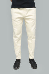 A person shown from the waist down wearing off-white slim-fit trousers, a black shirt, and black shoes against a solid gray background.
