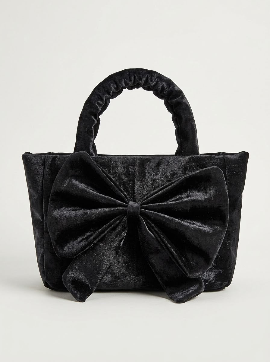 Front view of a black crushed velvet tote handbag featuring a large, decorative bow accent and ruched top handles against a light gray background.