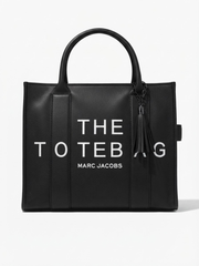 Front view of a structured black leather tote bag featuring white "THE TOTE BAG MARC JACOBS" typography and a matching tassel charm.