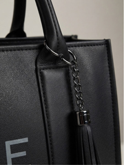 Close-up detail of the dark metal hardware ring, chain, and tassel attached to the handle base of the black leather tote bag.