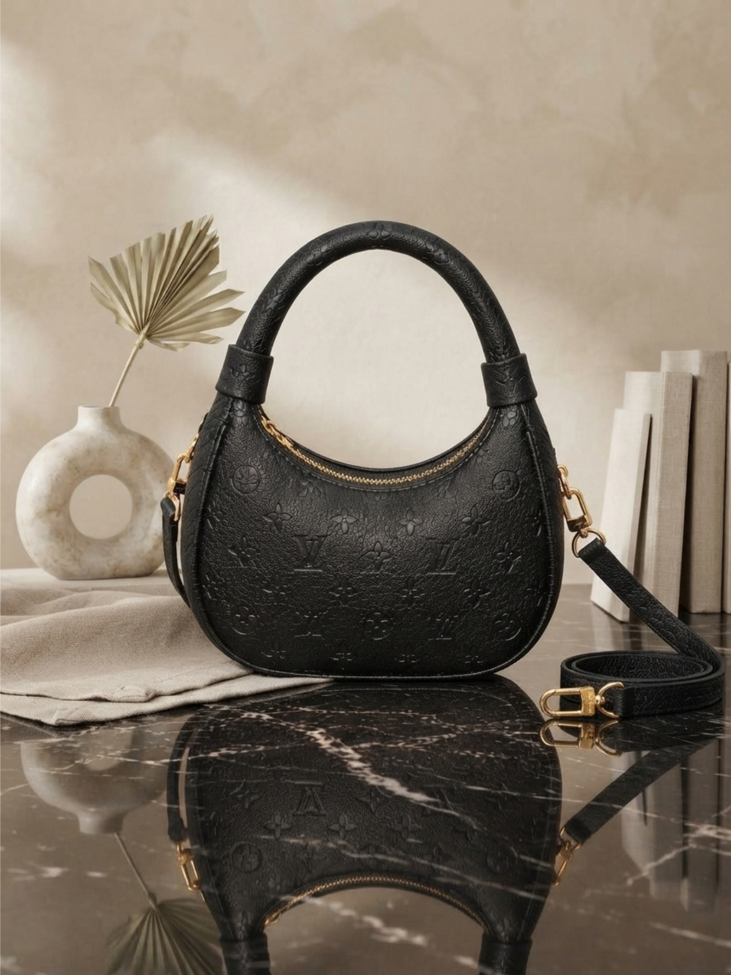 Front view of a black embossed Louis Vuitton Loop hobo handbag on a marble surface with a dried palm leaf vase and books in the background.