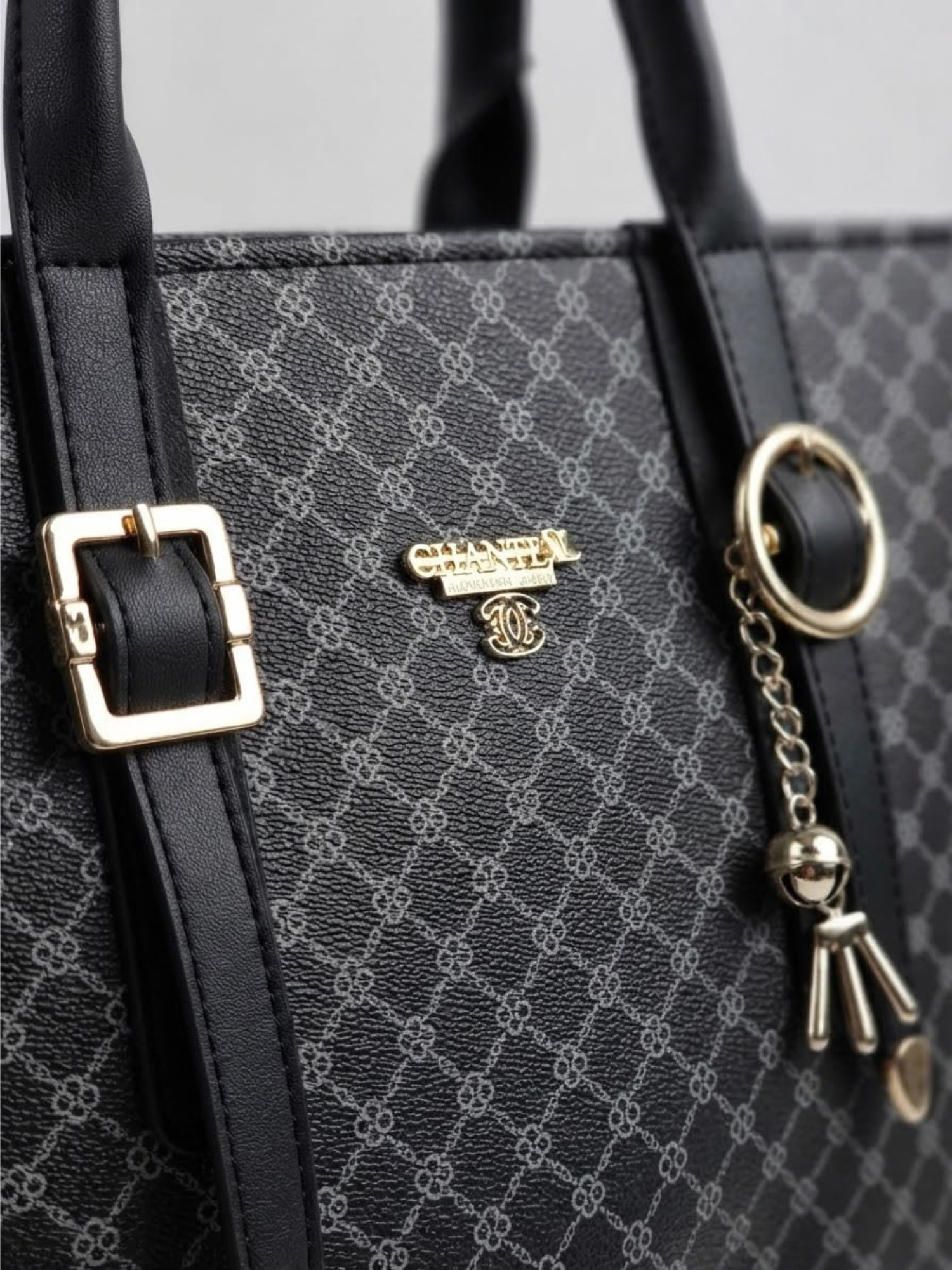 Close-up detail of the gold-tone buckle straps, brand logo badge, and charm keychain on the black patterned tote bag.