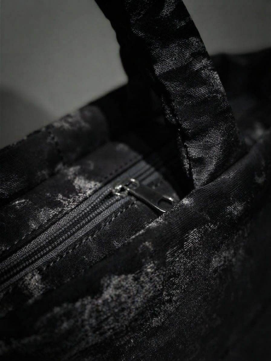 A detailed close-up photograph of the top zipper closure and silver-tone pull tab on the black abstract camo quilted handbag.