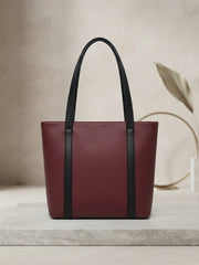 Front view of a maroon textured leather tote bag with black contrast handles resting on a marble surface.