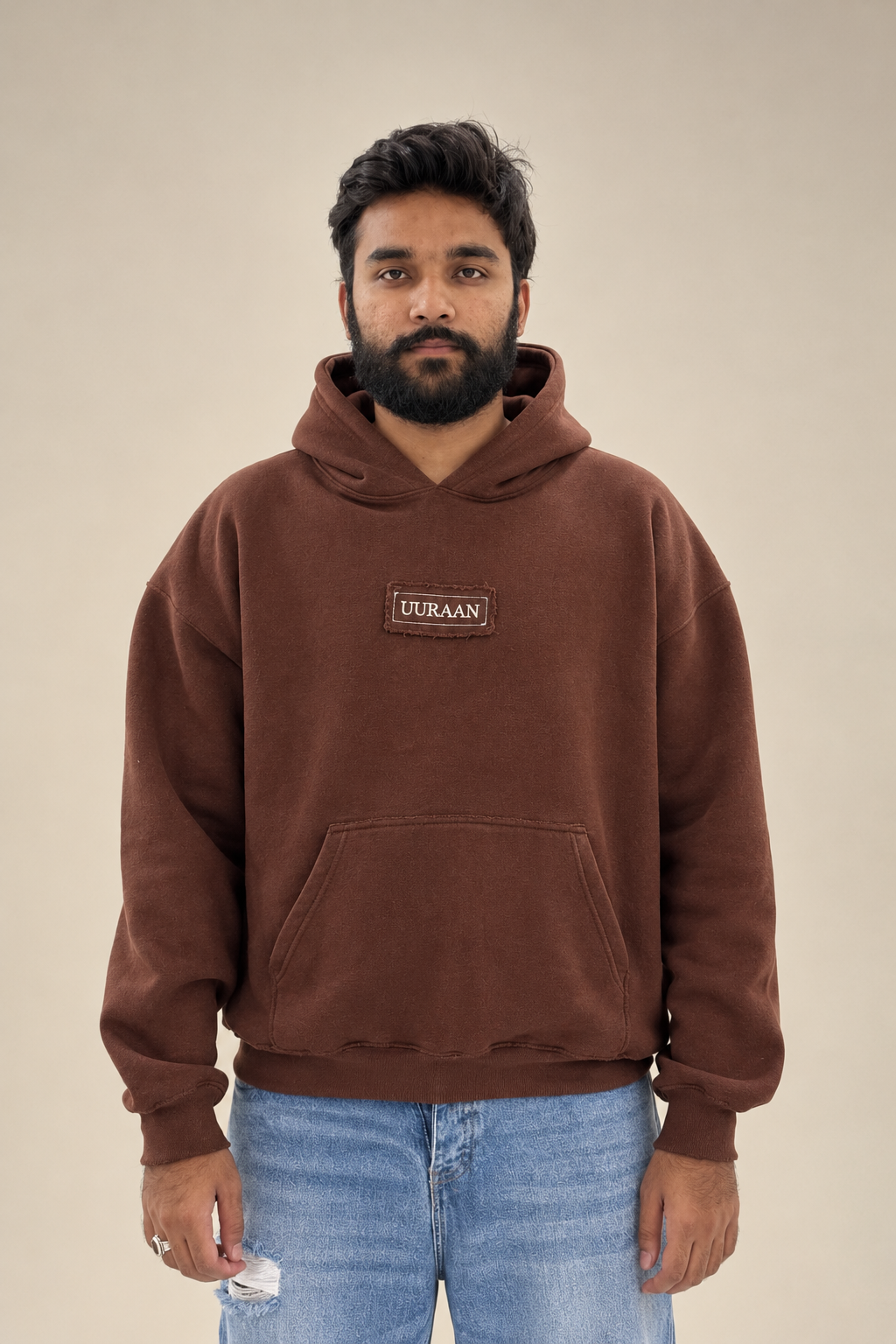 A male model wearing a premium brown fleece hoodie featuring a UURAAN logo chest patch and front kangaroo pocket paired with light blue jeans.
