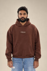A male model wearing a premium brown fleece hoodie featuring a UURAAN logo chest patch and front kangaroo pocket paired with light blue jeans.