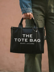 A person wearing a denim jacket and green trousers holding the black leather "THE TOTE BAG" by the top handles.