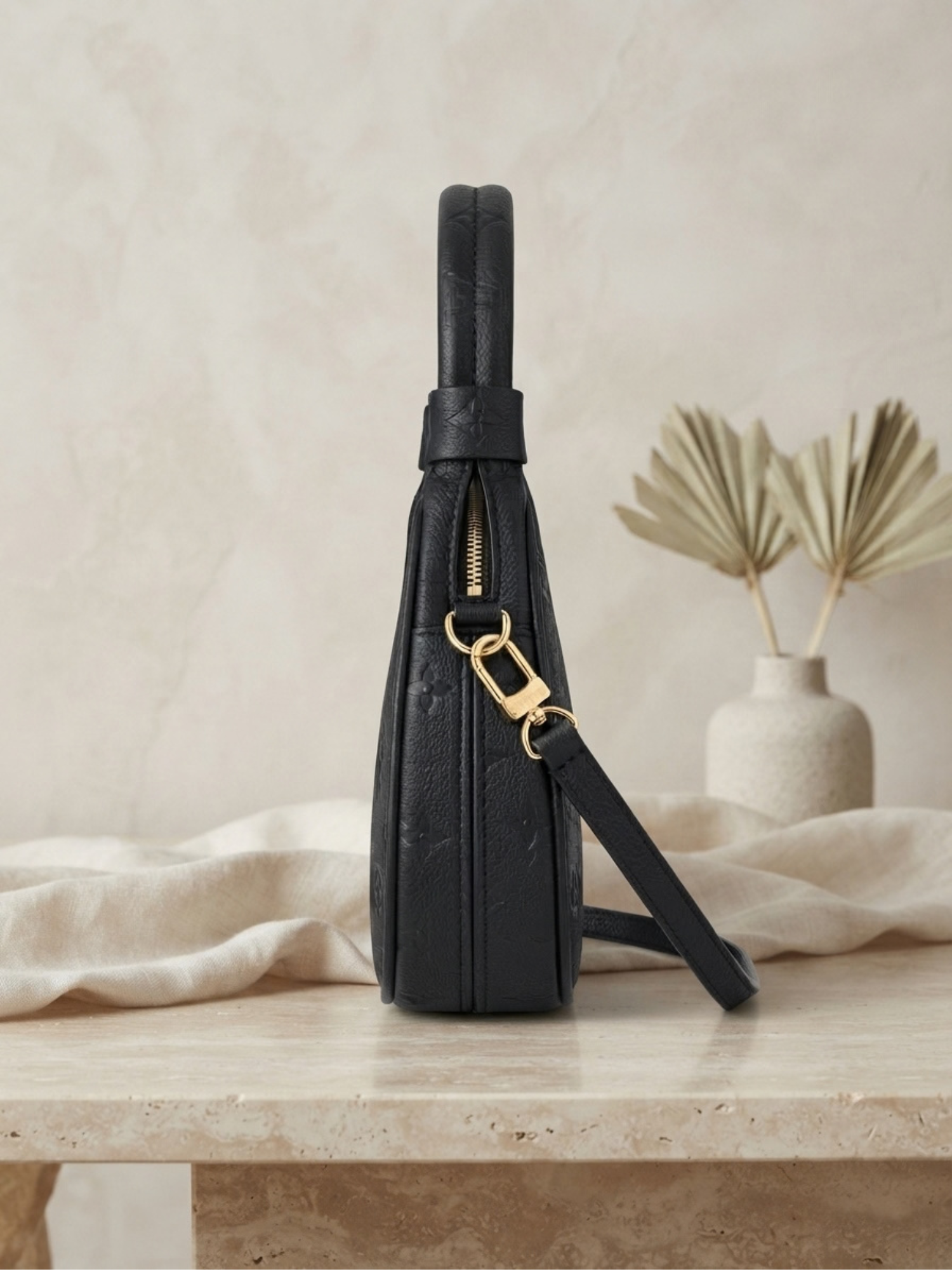 Side profile view of the black embossed Louis Vuitton Loop handbag on a light beige marble surface, showing the bag's depth and strap attachment.